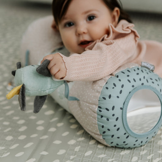 A baby in a pink sweater lies on a patterned play mat, holding a development toy attached to the Animal Adventures Tummy Time Pillow, a light blue, polka-dotted sensory baby pillow with a circular opening.