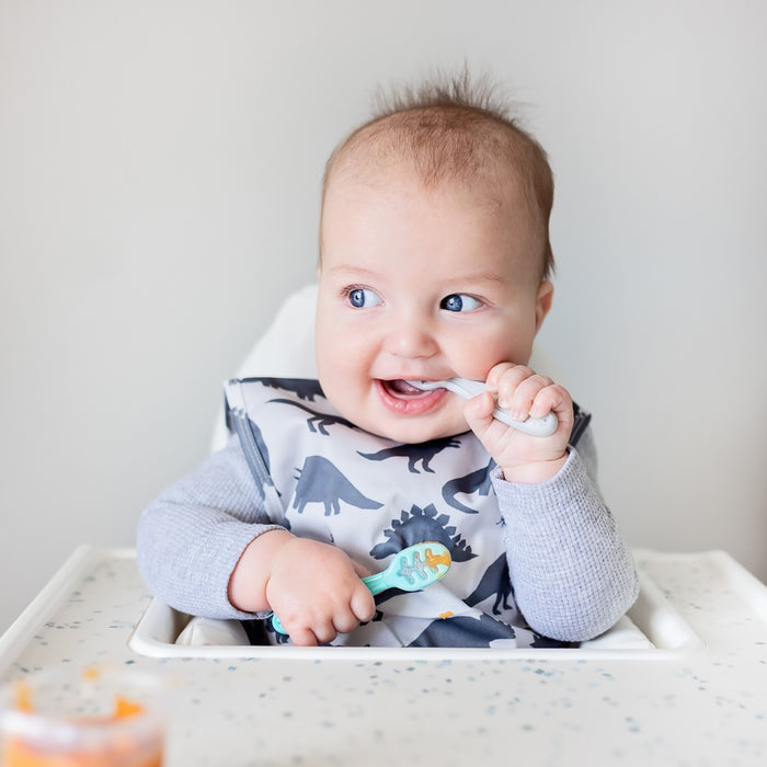 A baby in a highchair, wearing a dinosaur-patterned bib, smiles while holding a Nuby Baby's First Spoon in Aqua. Baby food on the tray is ideal for practicing with these self-feeding utensils in Peach and Light Blue.