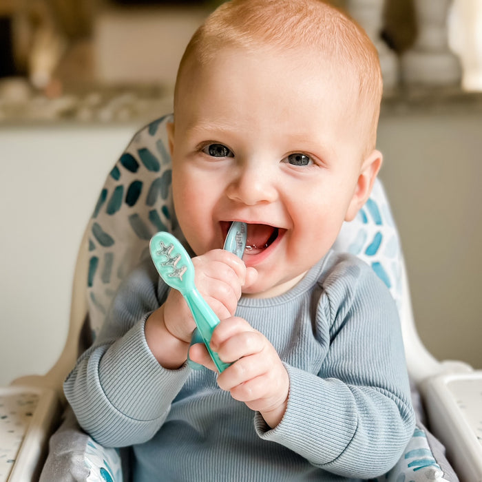 A smiling baby with light hair in a high chair happily chews on Nuby Baby's First Spoons in Aqua, Peach, and Light Blue while wearing a light blue long-sleeve shirt and enjoys exploring these self-feeding utensils.