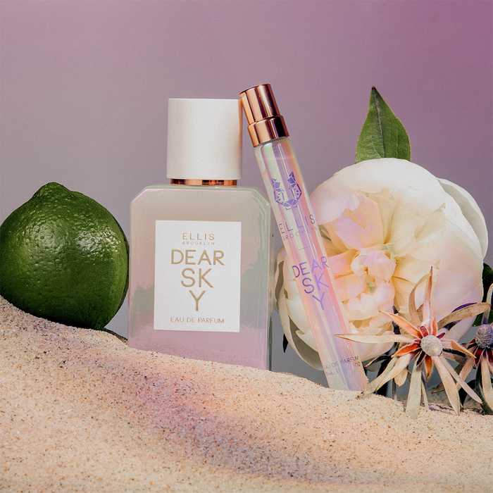 Dear Sky 50ml Eau De Parfum and Dear Sky 10ml Eau De Parfum Travel spray in sand next to Bergamot and flowers, against purple background.