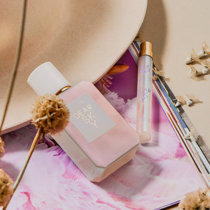 Dear Sky 50ml Eau De Parfum and Dear Sky 10ml Eau De Parfum Travel spray resting on purple and white magazine image and beige hat with dried flowers, on beige background.
