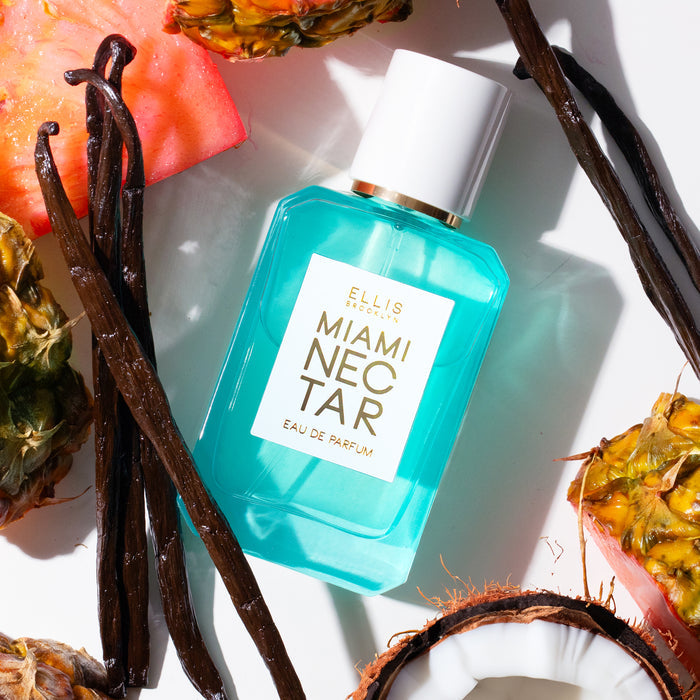 MIAMI NECTAR Eau de Parfum 50ml lays next to slices of pink pineapple, a cut open coconut, and vanilla beans on a white background