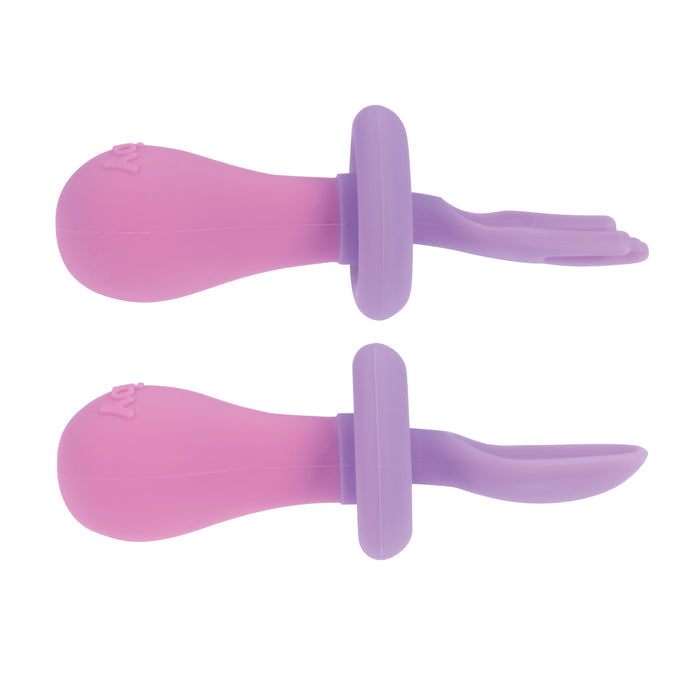 The First Fork & Spoon Set includes two silicone sets in pink and purple, each with rounded handles and safety guards—one standard tip spoon and one split-tip fork—perfect self-feeding utensils for babies.