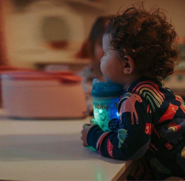 A young child in vibrant rainbow and unicorn pajamas sits at a table holding a glowing green Nuby Insulated Light-Up Easy Sip Cup with a No Spill, bite-resistant spout, keeping drinks cool in the softly blurred indoor setting.