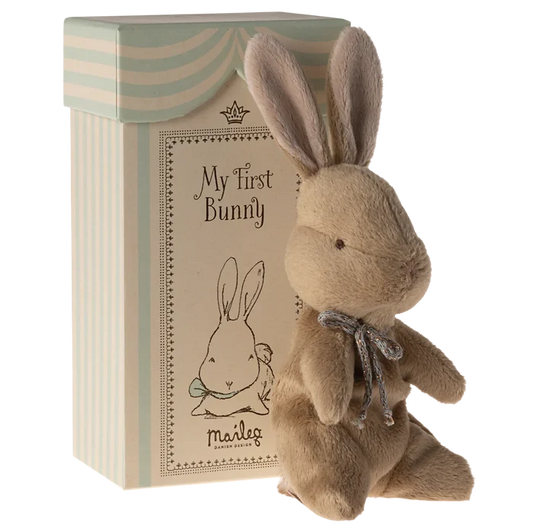 A plush toy rabbit with a brown body, beige ears, and a blue ribbon around its neck, sitting next to its beige and blue striped gift box with 'My First Bunny' text on it.