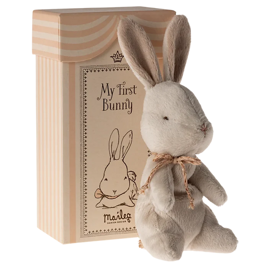 A soft plush toy rabbit with an off-white color, sitting next to its beige cardboard gift box which has 'My First Bunny' written on it.