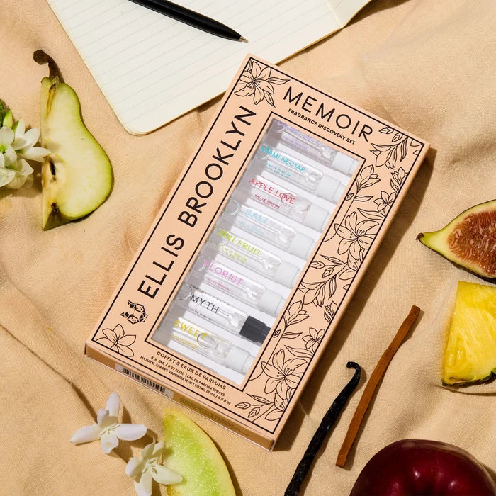Memoir Fragrance Discovery Set box with beige and black floral design showing Dear Sky Eau De Parfum 1.5ml Vial, Miami Nectar Eau De Parfum 1.5ml Vial, Apple Love Eau De Parfum 1.5ml Vial, Salt Eau De Parfum 1.5ml Vial, Sun Fruit Eau De Parfum 1.5ml Vial, Florist Eau De Parfum 1.5ml Vial, Myth Eau De Parfum 1.5ml Vial, Sweet Eau De Parfum 1.5ml Vial, and Vanilla Milk Eau De Parfum 1.5ml Vial, on tan fabric with fig, pineapple, apple, sandalwood, vanilla bean, pear, white flowers, and notebook wi