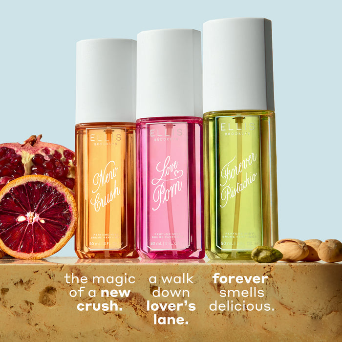 NEW CRUSH Perfume Mist 90ml, LOVE POM Perfume Mist 90ml, and FOREVER PISTACHIO Perfume Mist 90ml on tan marble between sliced pomegranate, blood orange, and pistachios against a light blue background with white text that reads "the new magic of a new crush. a walk down lover's lane. forever smells delicious."